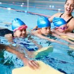Female Coach In Water Giving Group Of Children Swimming Lesson In Indoor Pool